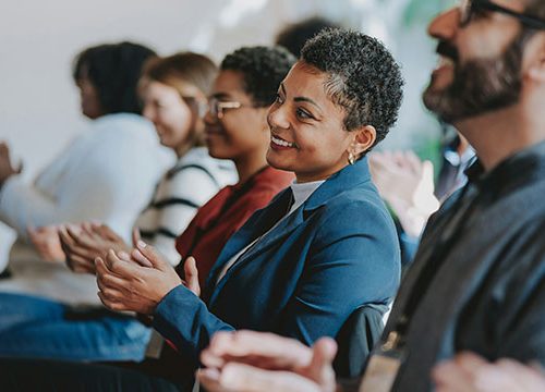 A man and three woman sat on the front row clapping a the speaker at a professional event.