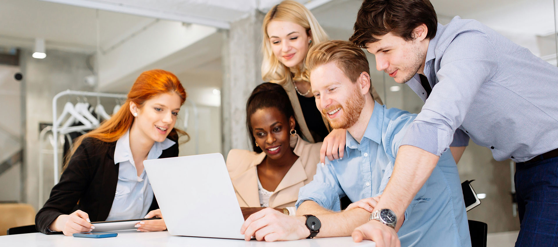 Mixed group of men and woman gathered around a laptop in an office, smiling and talking.