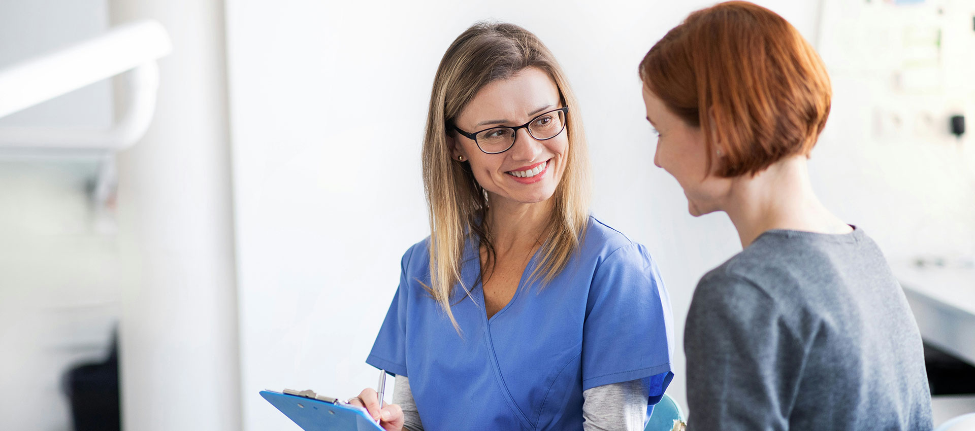 Female, blonde nurse with a clipboard in her hands talking to her female patient with short read hair.