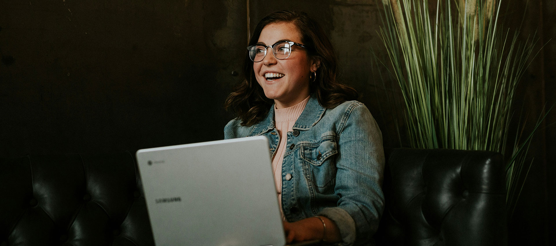 A young woman wearing glasses and smiling, sat on a black leather sofa with a laptop in her lap.