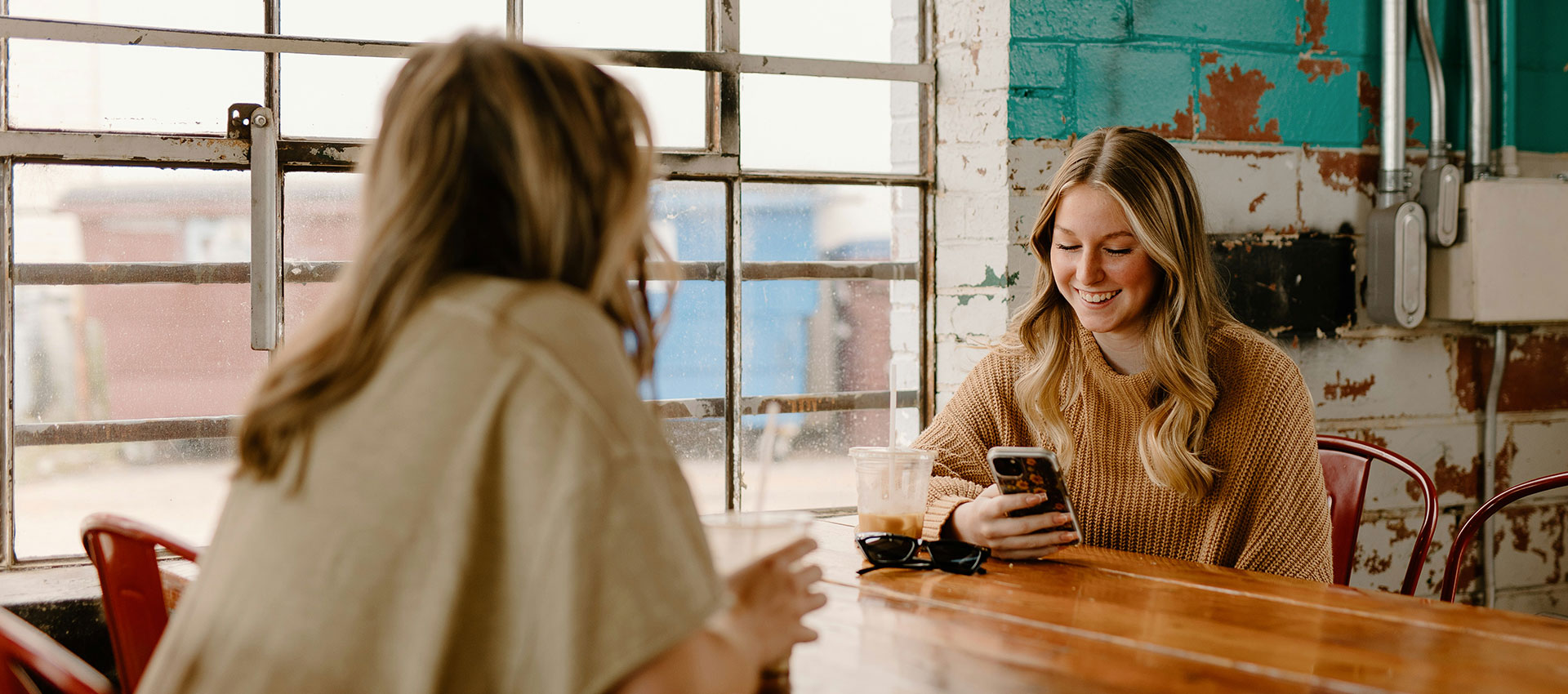 Two blonde woman sat a wooden table in a cafe, chatting and looking at their phones.
