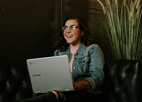 A young woman wearing glasses and smiling, sat on a black leather sofa with a laptop in her lap.