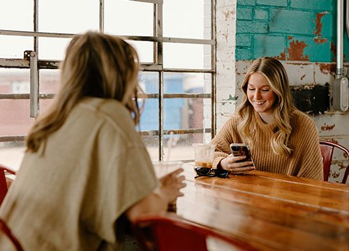 Two blonde woman sat a wooden table in a cafe, chatting and looking at their phones.