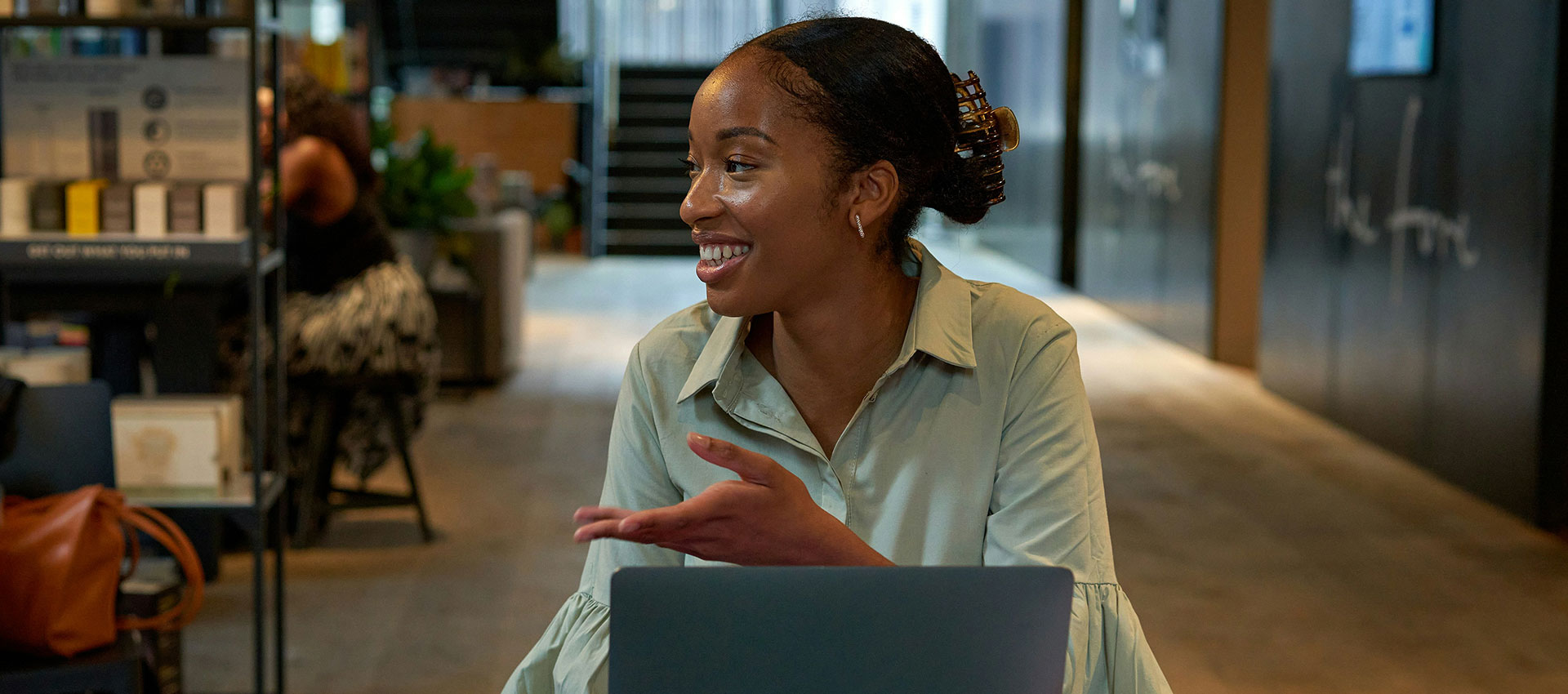A young black woman sat at her desk in a modern office talking to someone off camera.
