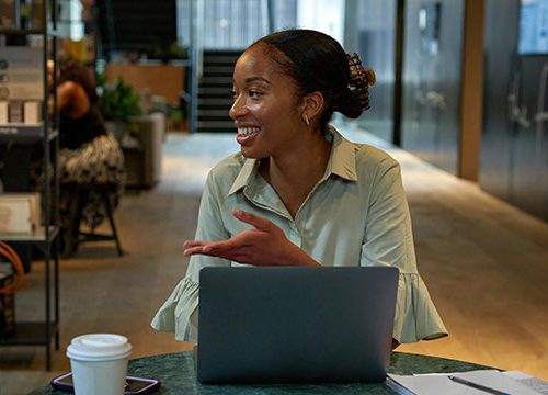 A young black woman sat at her desk in a modern office talking to someone off camera.