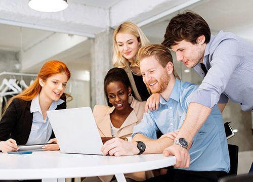 Mixed group of men and woman gathered around a laptop in an office, smiling and talking.