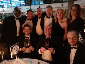 A group of people dress in black tie dresses and tuxedos gather round a table at an awards ceremony smiling for the camera.