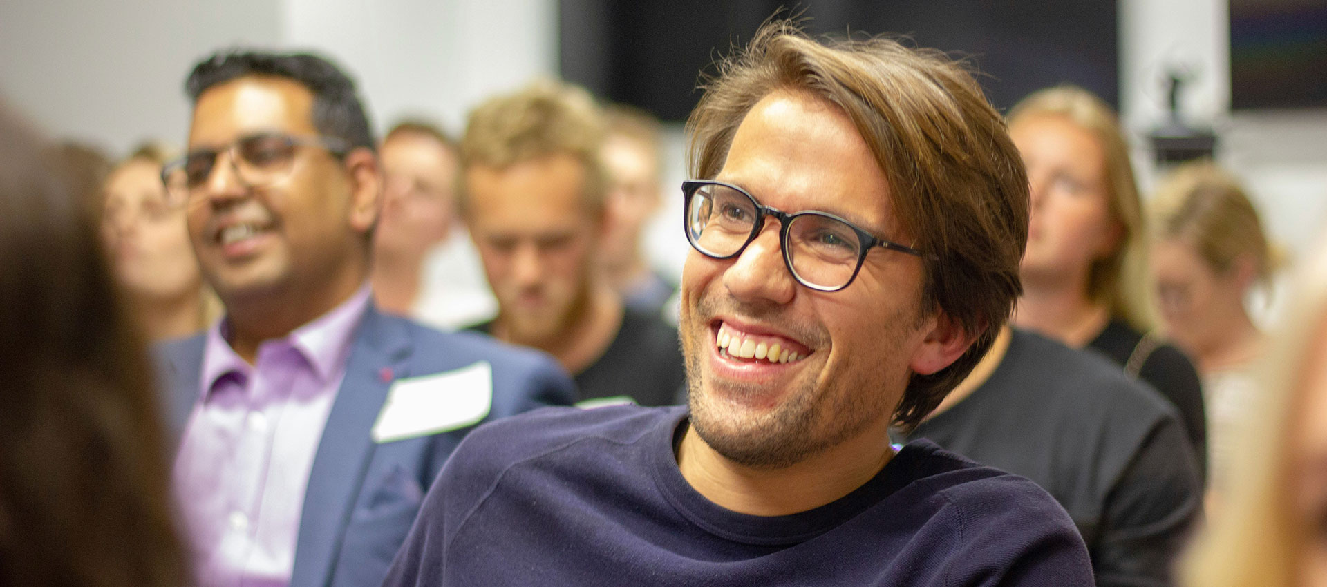 Close up of a middle aged man wearing glasses and a navy jumper, laughing whilst sat in the audience of an exhibition talk.