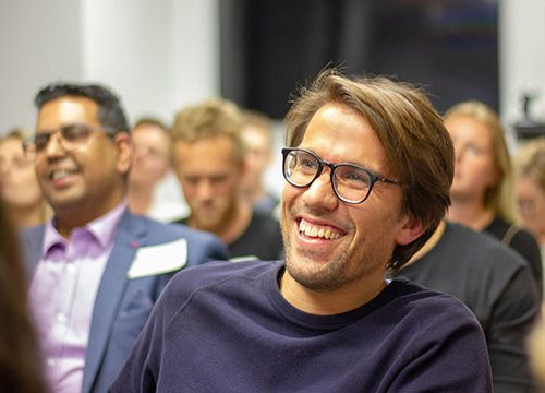 Close up of a middle aged man wearing glasses and a navy jumper, laughing whilst sat in the audience of an exhibition talk.