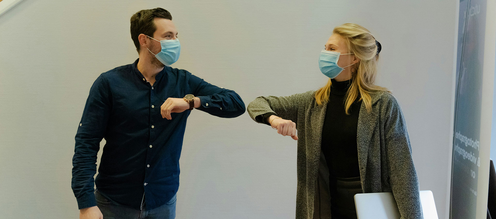 A middle aged man in a navy shirt and a blonde woman in a grey cardigan touching elbows in greeting while wearing surgical masks covering their mouths.