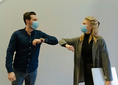 A middle aged man in a navy shirt and a blonde woman in a grey cardigan touching elbows in greeting while wearing surgical masks covering their mouths.