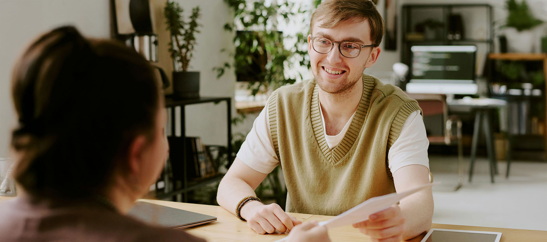 Young man wearing glasses and a green jumper sat at a desk in a modern office, passing a piece of paper to a girl with dark hair.