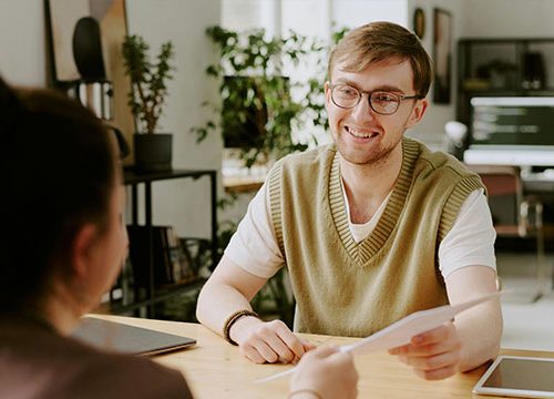 Young man wearing glasses and a green jumper sat at a desk in a modern office, passing a piece of paper to a girl with dark hair.