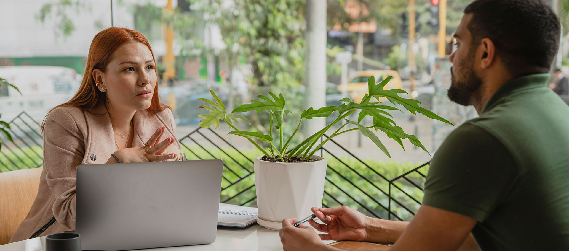 A young woman with red hair sat a desk with a laptop in front of her talking to a man wearing a green polo top with dark facial hair sat across the table from her.