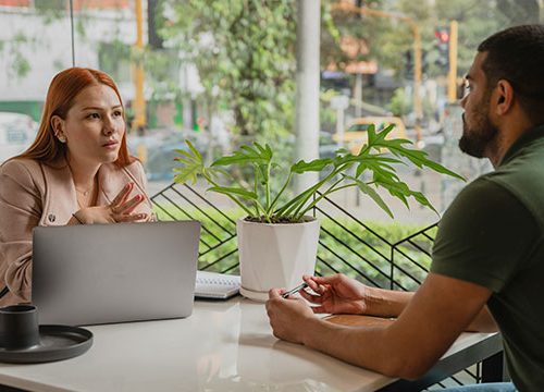 A young woman with red hair sat a desk with a laptop in front of her talking to a man wearing a green polo top with dark facial hair sat across the table from her.