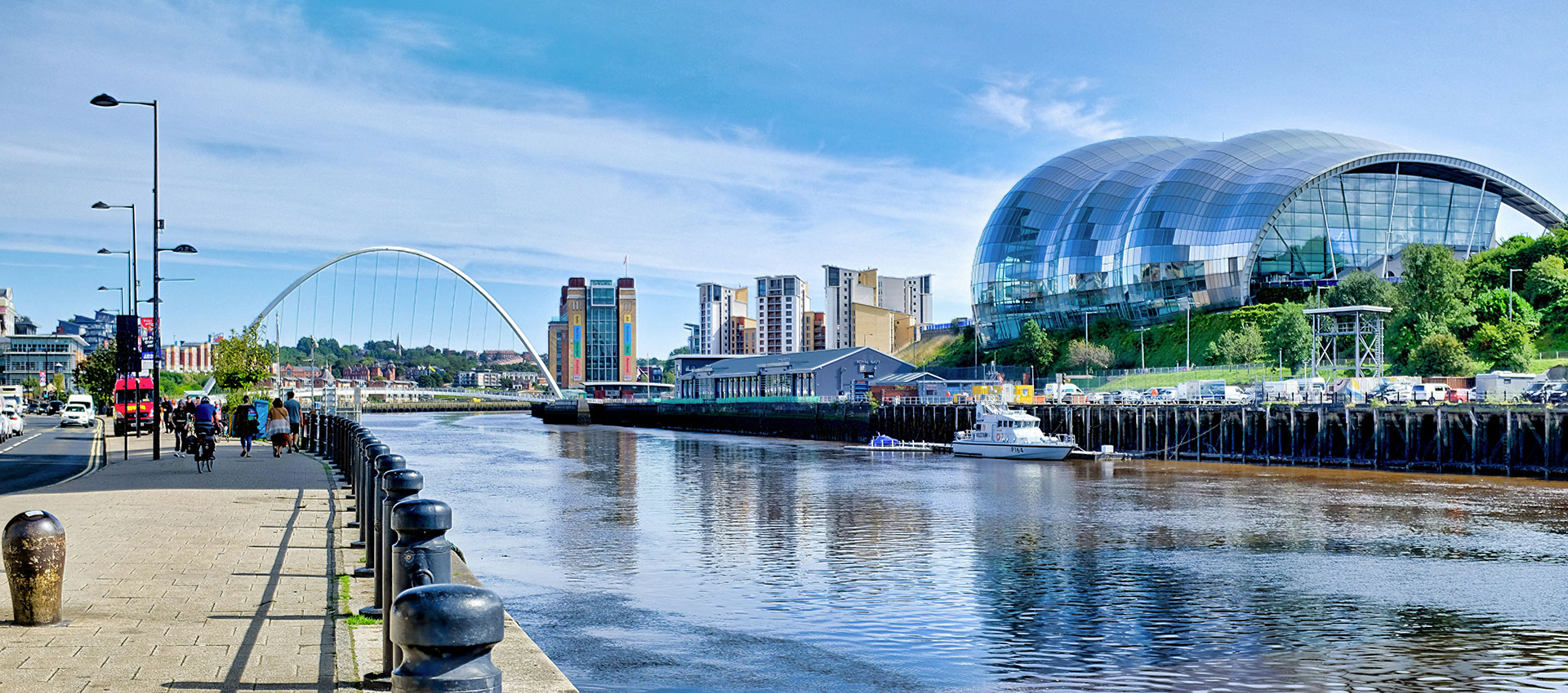 People walking down the pavement, the Millenium bridge, the Baltic arts centre and the Sage Museum on Newcastle's quayside photographed on a day with blue sunny skies.