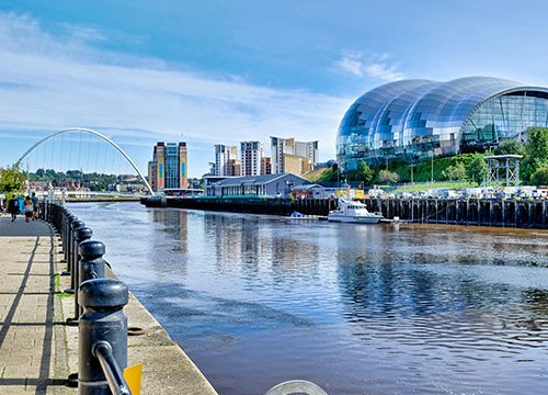 People walking down the pavement, the Millenium bridge, the Baltic arts centre and the Sage Museum on Newcastle's quayside photographed on a day with blue sunny skies.