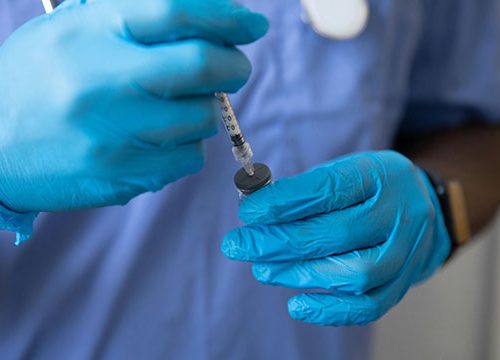Close up of a black male nurse wearing surgical gloves and holding a syringe.