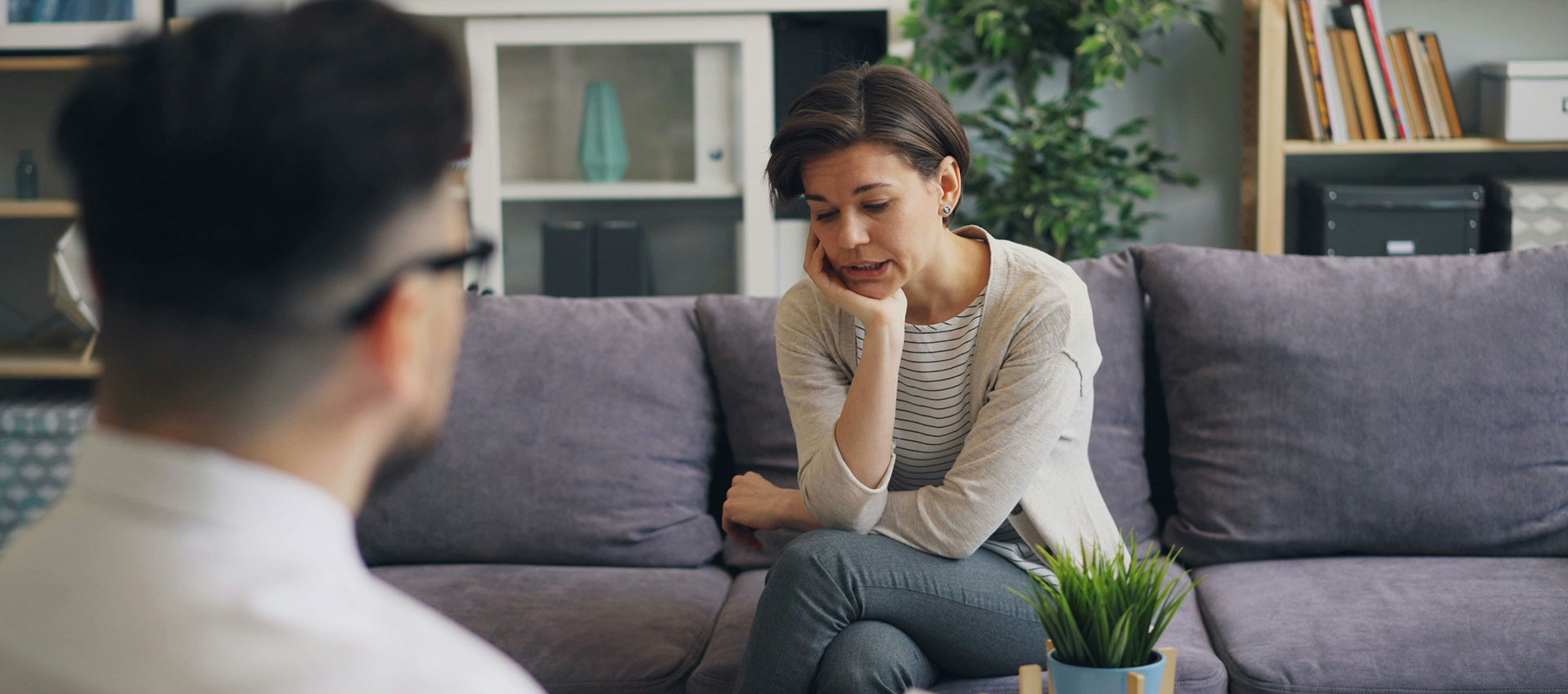 Middle aged woman with short dark hair sat crossed legged on a purple sofa with her elbow on her knee and her chin resting on the palm of her hand.