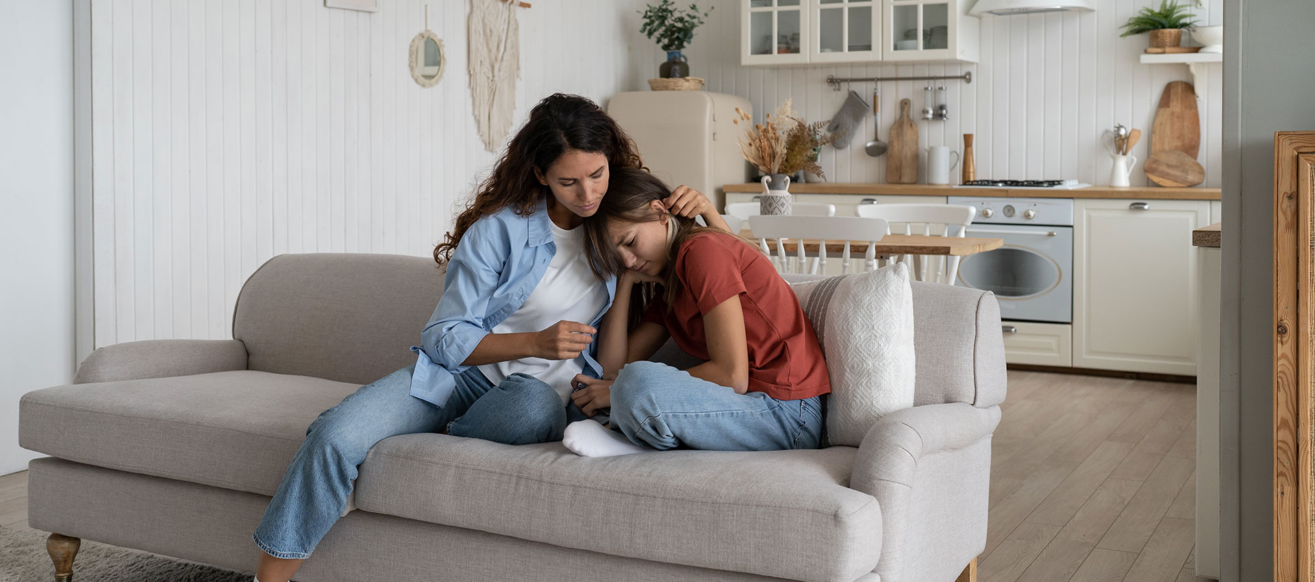A woman with long brown hair sat on a cream sofa in an open plan kitchen-living room, cuddling and comforting a young girl in a red top.