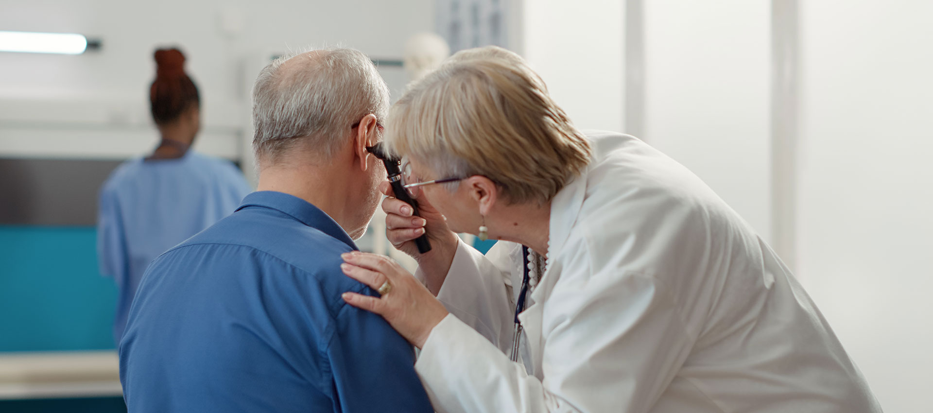 An older female health professional bending down towards an older mans ear with a temperature taker.
