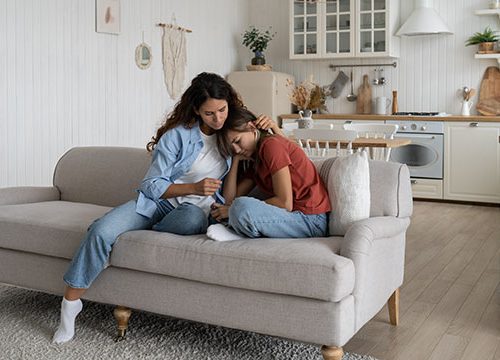 A woman with long brown hair sat on a cream sofa in an open plan kitchen-living room, cuddling and comforting a young girl in a red top.