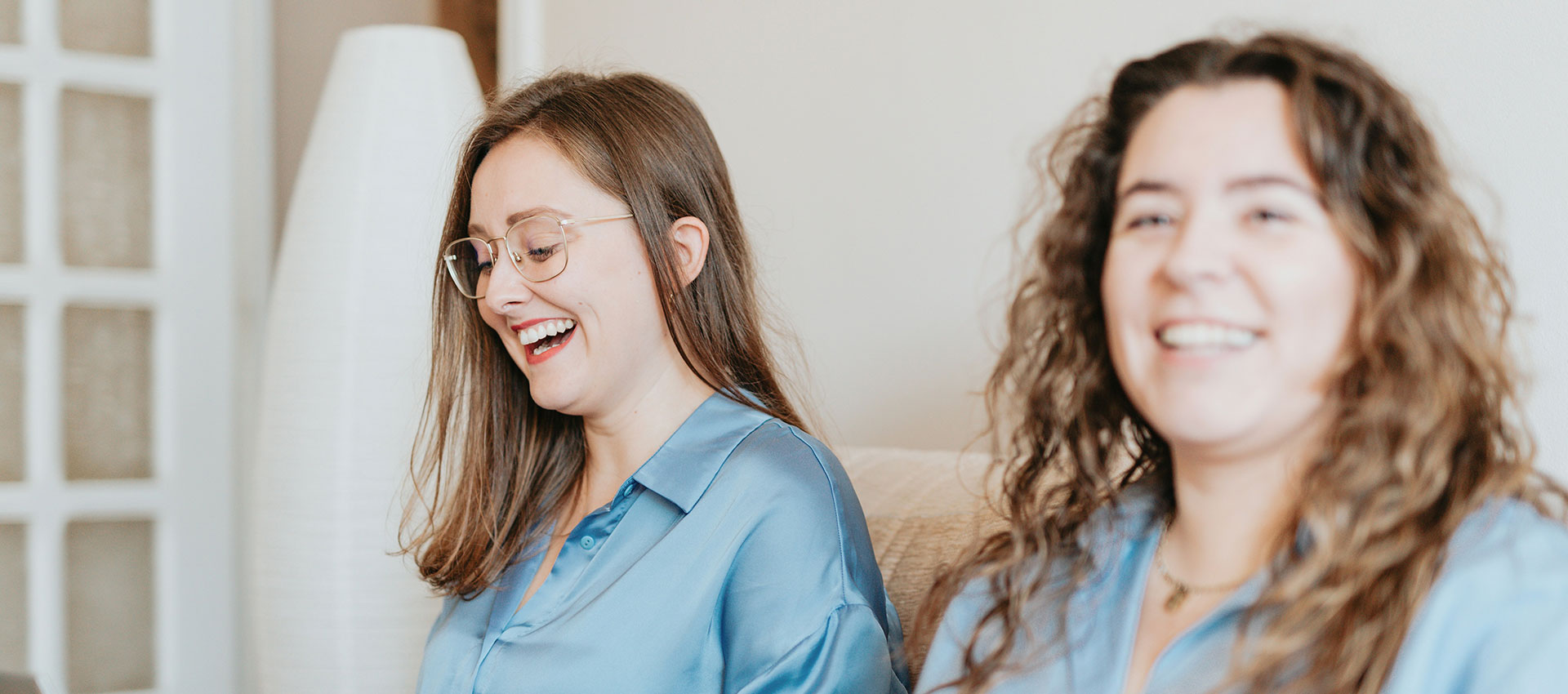 Two women in blue satin shirts sit on a sofa with laptops on their laps, laughing.