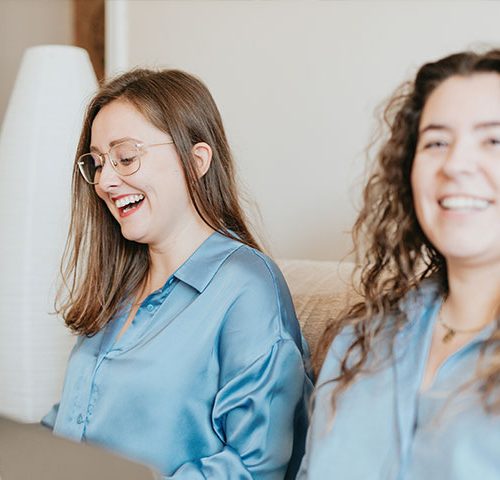 Two women in blue satin shirts sit on a sofa with laptops on their laps, laughing.