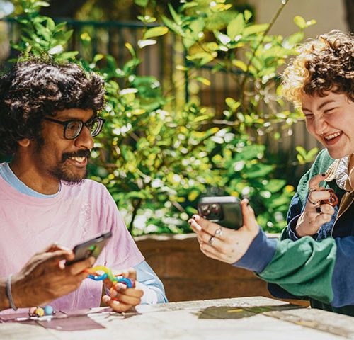 An asian man in a pink and blue top sits outside laughing with a woman with curly hair and a blue and green striped polo top on.