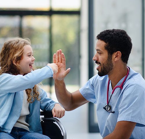A male nurse with dark hair and dark facial hair squats down next to a little girl in a wheelchair to give her a high five.