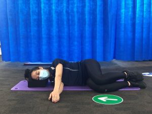 Person lies on a purple exercise mat, raising one arm vertically, wearing a mask. They rest their head on a block. A blue curtain serves as the backdrop, with directional floor signage.