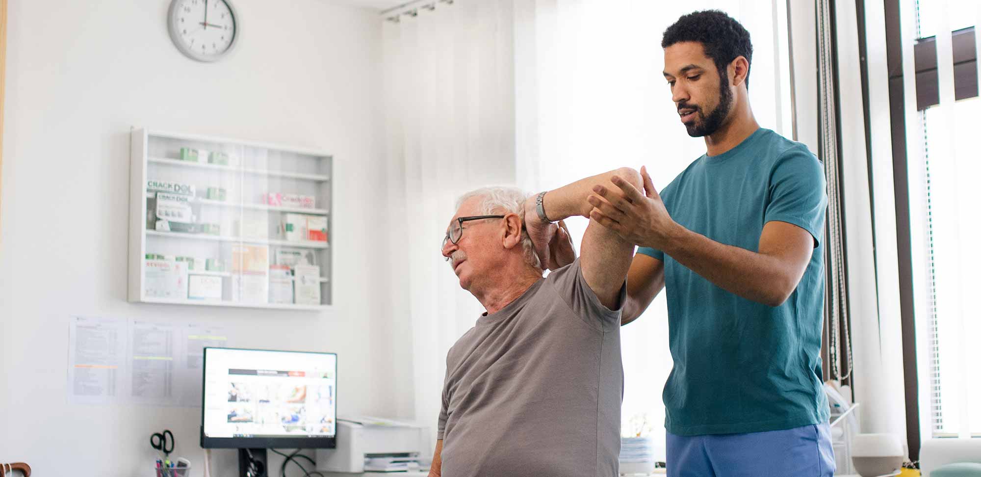 A healthcare professional assists an elderly man with arm exercises in a bright clinic room. A wall clock, medicine cabinet, and computer monitor are visible in the background.