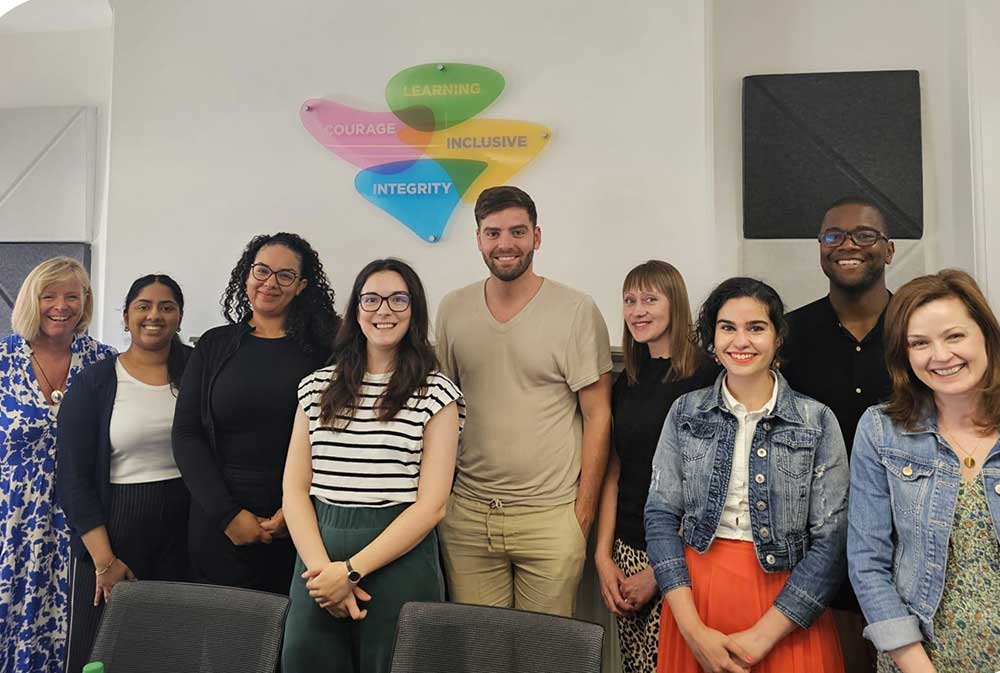 A group of nine people stands smiling in an office. Behind them, a colorful wall decoration displays the words 