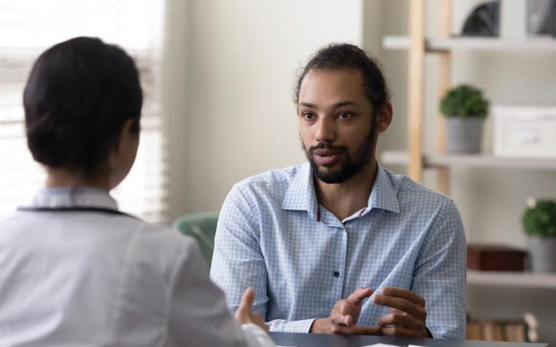 A healthcare professional and a patient are seated and conversing in a well-lit office with shelves containing books and plants, emphasizing a professional healthcare consultation environment.