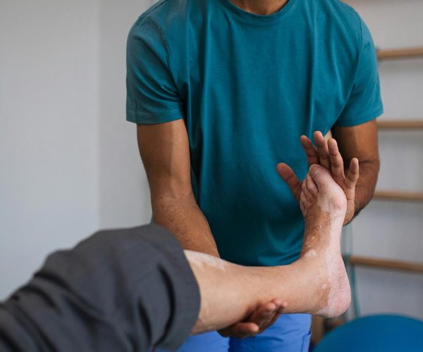 A therapist in a teal shirt assists a client by supporting their foot and ankle during a physical therapy session, with exercise equipment in the background.