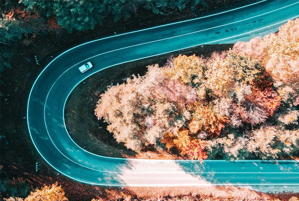 A white car navigates a sharp, winding road surrounded by vibrant autumn-colored trees in a forested area, viewed from above.