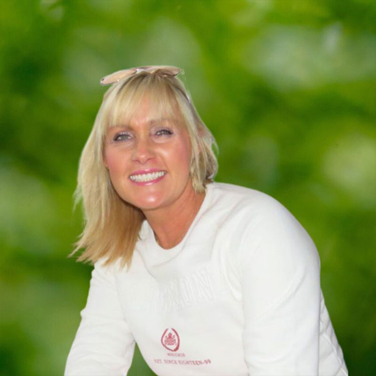 A person is smiling while sitting in a relaxed outdoor setting with lush green foliage in the background. They are wearing a white top.