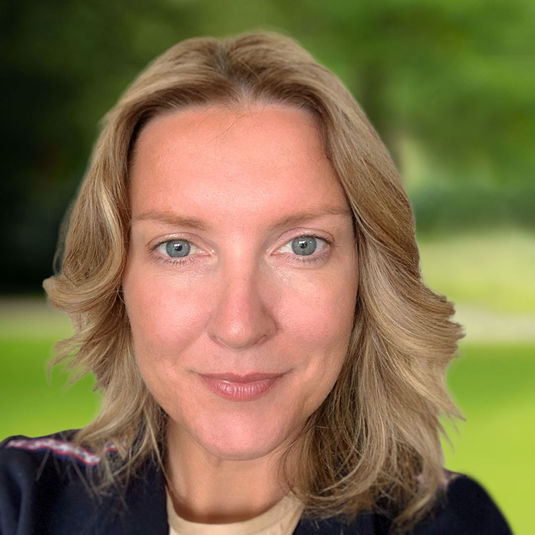A person with light hair is looking directly at the camera, smiling faintly. They are outdoors with a blurred background of greenery, suggesting a park or garden setting.