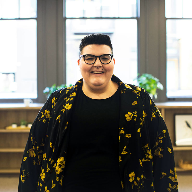 A woman with short dark hair wears a black top and a black and yellow patterned floral blazer smiles at the camera while stood in an office.