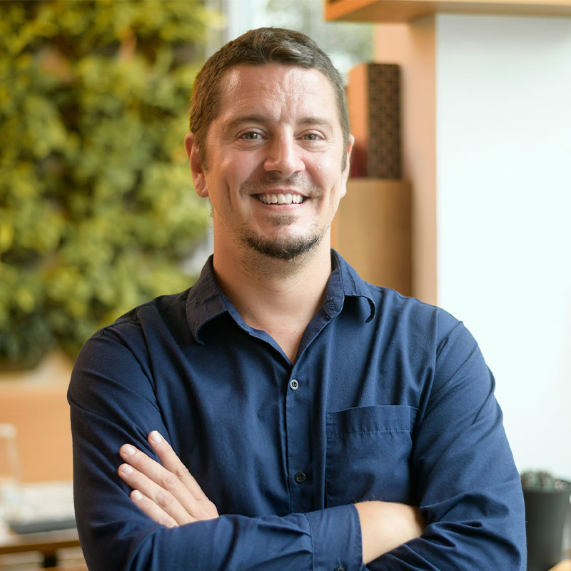A man wearing a blue shirt stands with his arms crossed over his chest as he smiles at the camera.