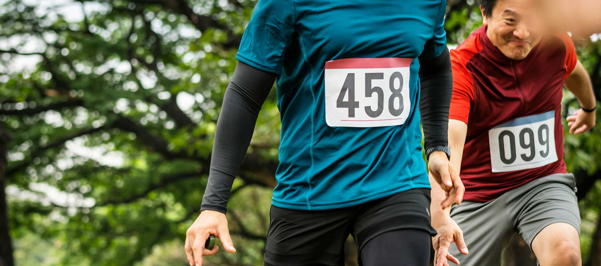 Close up of two men running in a race, the man on the left wears black skins and a blue top with the race number 458 stuck to his top and the man on the right wears grey shorts and a red top with the number 099 stuck to his top.