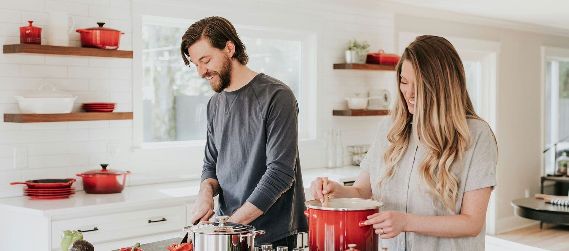 A man and a woman cook together in a bright kitchen, using red cookware on a counter. Shelves in the background hold additional red kitchen items.