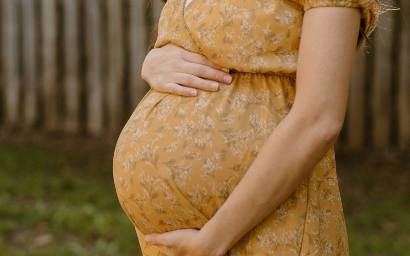 A person in a yellow patterned dress stands in a grassy area, gently holding their pregnant belly with both hands. A wooden fence is visible in the background.