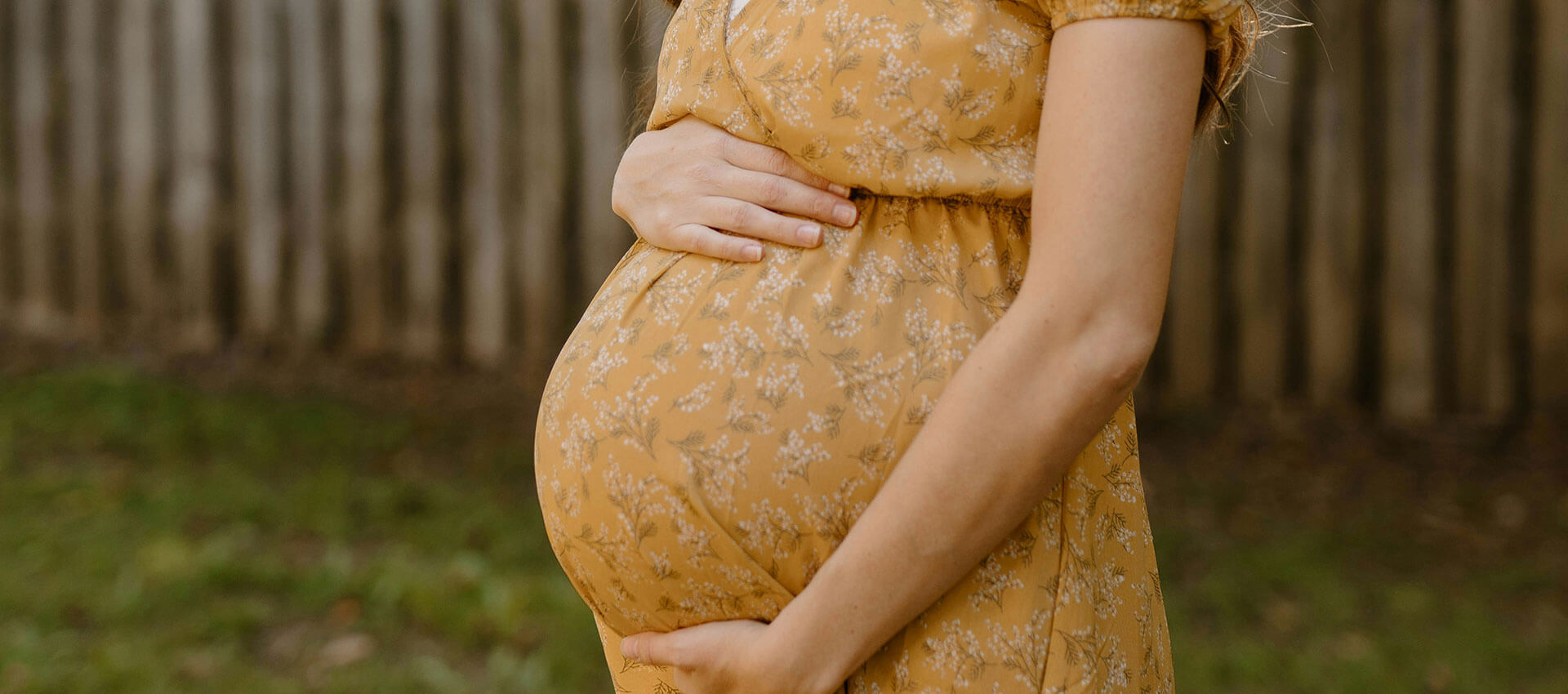 A person in a yellow patterned dress stands in a grassy area, gently holding their pregnant belly with both hands. A wooden fence is visible in the background.