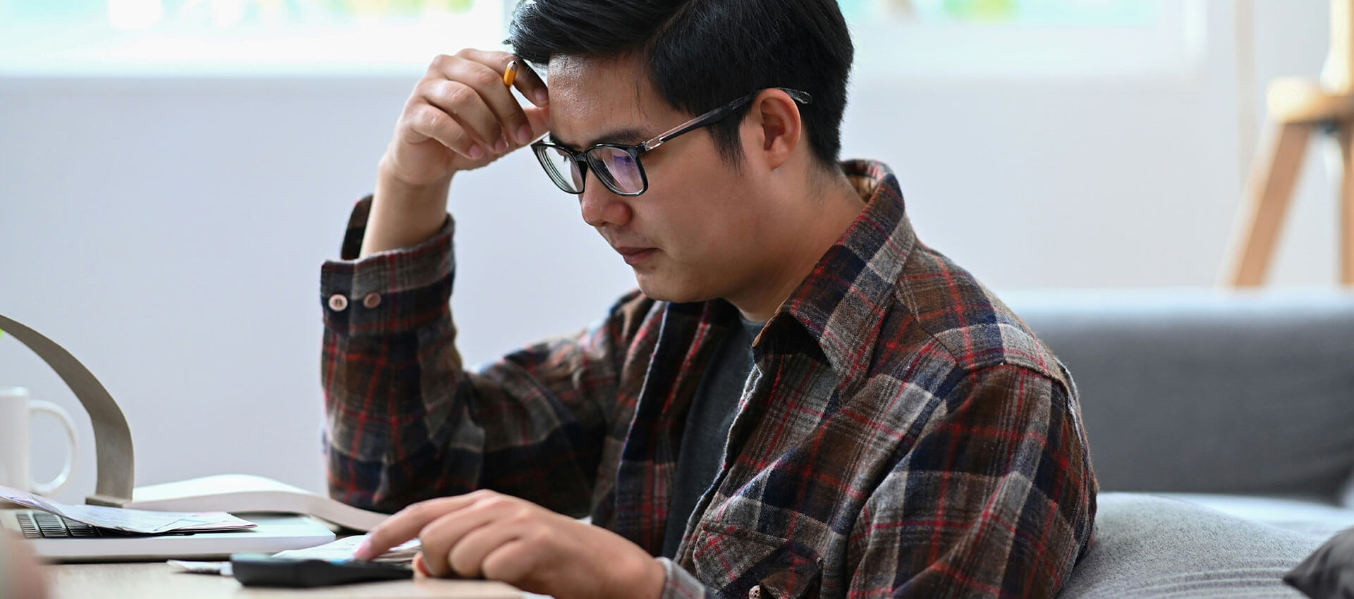 A man in a plaid shirt sits at a desk, focused on a tablet, with a pen held to his temple. A laptop and mug are nearby, suggesting a work environment.