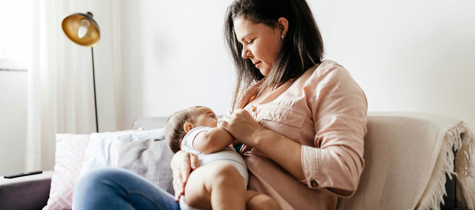 A person feeds a baby with a bottle while sitting on a beige couch. A floor lamp and a small table with decorations are in the background.