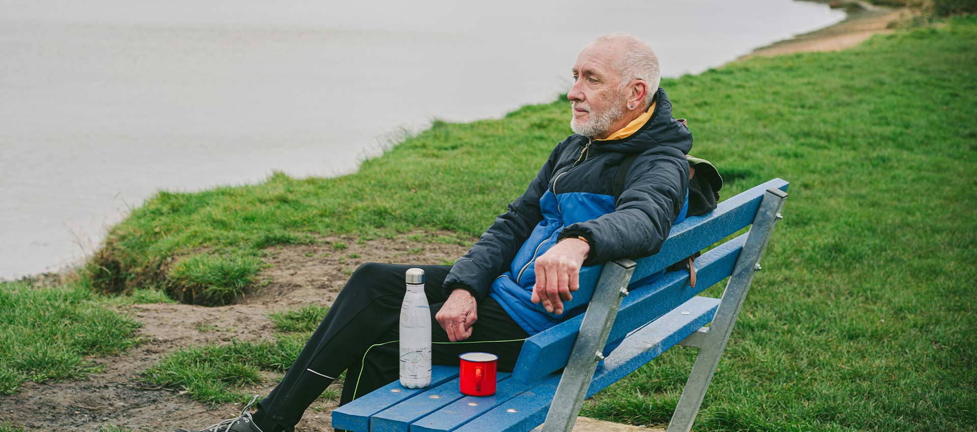 A older man with grey hair and beard in walking gear, sat crossed leg on a metal bench staring out over the the water in front of him.