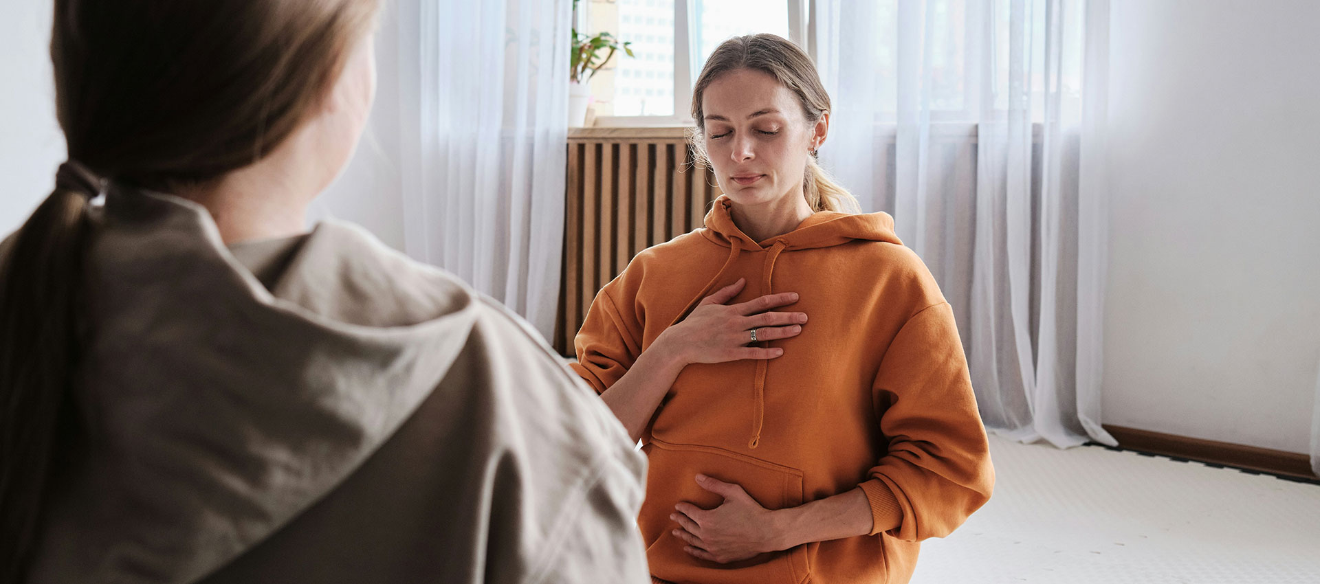 A woman with blonde hair wearing an orange hoody sits on the floor with her eyes closed and one hand on her chest and one hand on her stomach opposite another woman in a beige hoody as they practise breathing exercises.