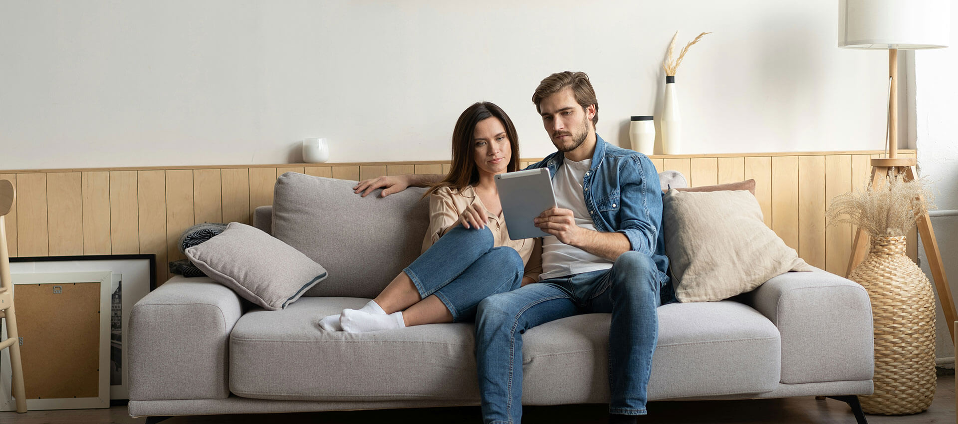 A couple sits closely on a beige sofa, engaged in viewing a tablet. They are in a cozy living room with neutral tones, decorative cushions, and a tall lamp beside them.