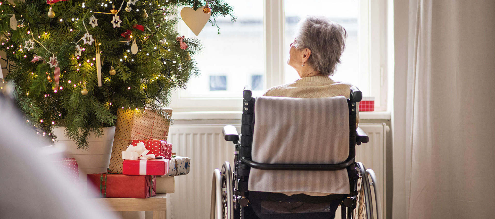 An older woman with grey hair sat in a black wheel chair looking out her window, next to a Christmas tree with lots of wrapped presents underneath.
